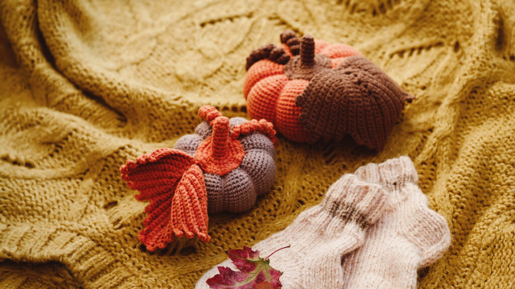 Crocheted pumpkins and cozy socks rest on a yellow knitted blanket with a red autumn leaf nearby.