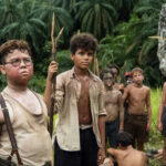 A group of dirty, shirtless boys stand in tall grass in front of palm trees, looking serious and alert.