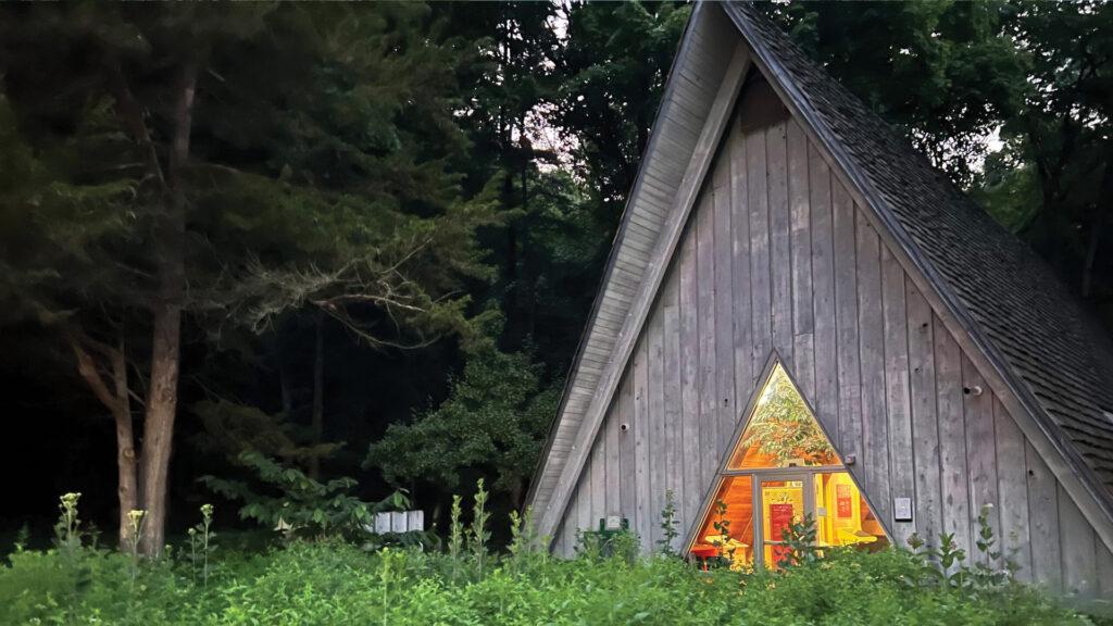 A-frame cabin with warm light inside, surrounded by trees and grass in a forest setting.