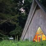 A-frame cabin with warm light inside, surrounded by trees and grass in a forest setting.