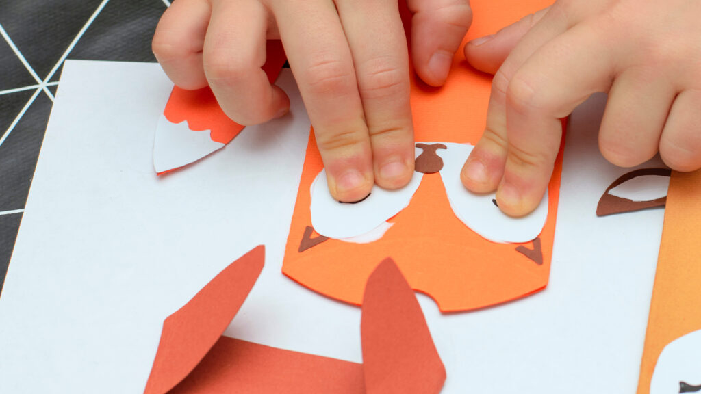 Child’s hands assembling an orange paper animal face with white eyes, likely a fox, on a table.