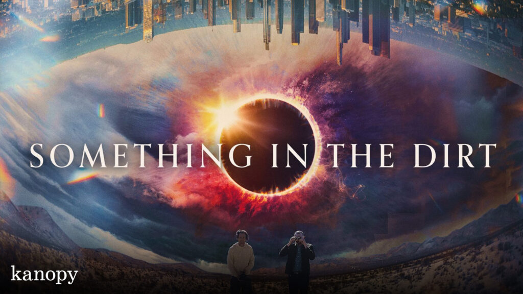 Two men stand in a desert beneath a dramatic, swirling sky, an eye-like burst at the center. Text reads “Something in the Dirt.