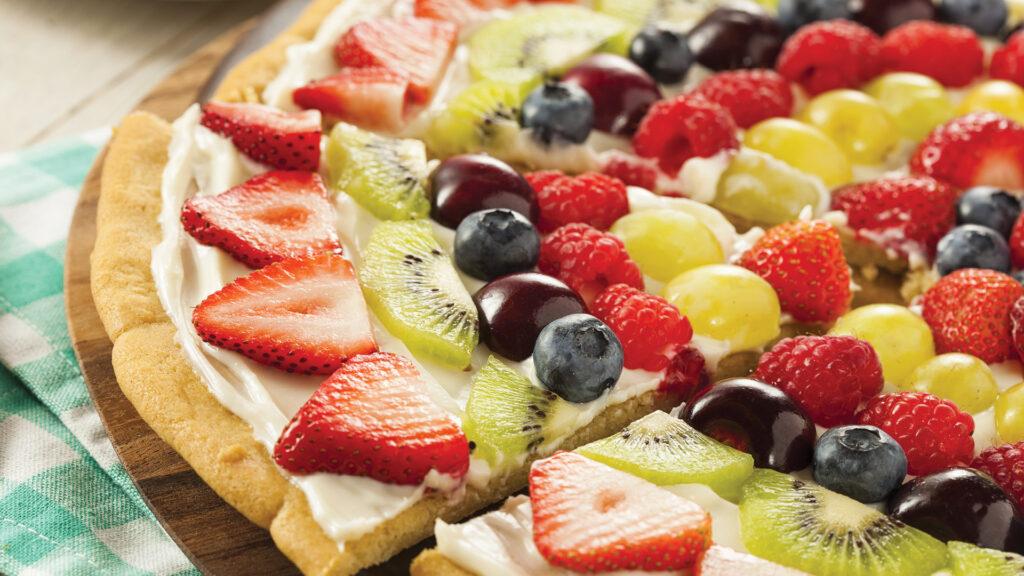 Colorful fruit pizza with strawberries, kiwi, grapes, blueberries, and raspberries on a cream-covered crust.