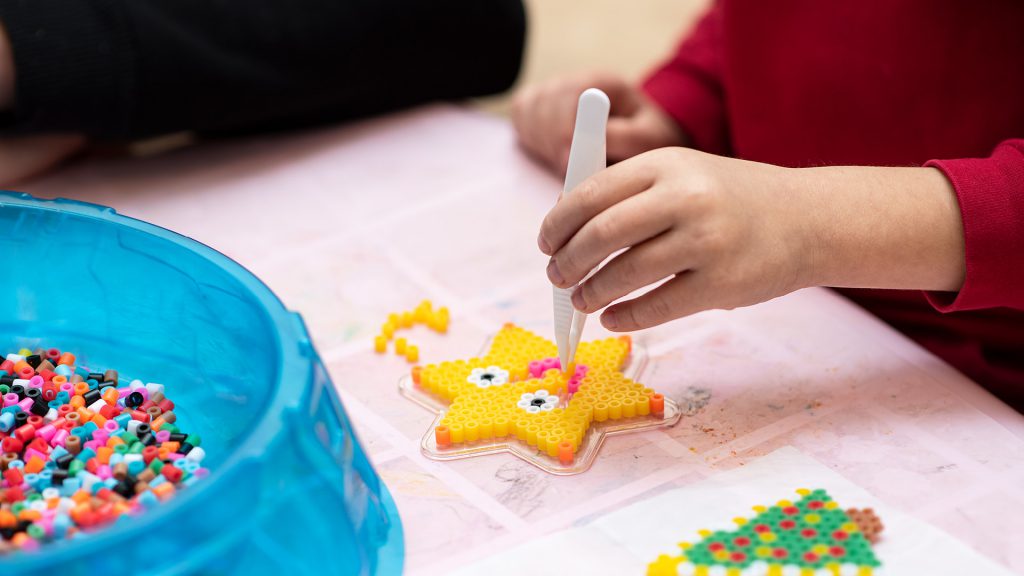 Child arranging colorful beads into a star shape using a tweezer, with a blue container of beads nearby.