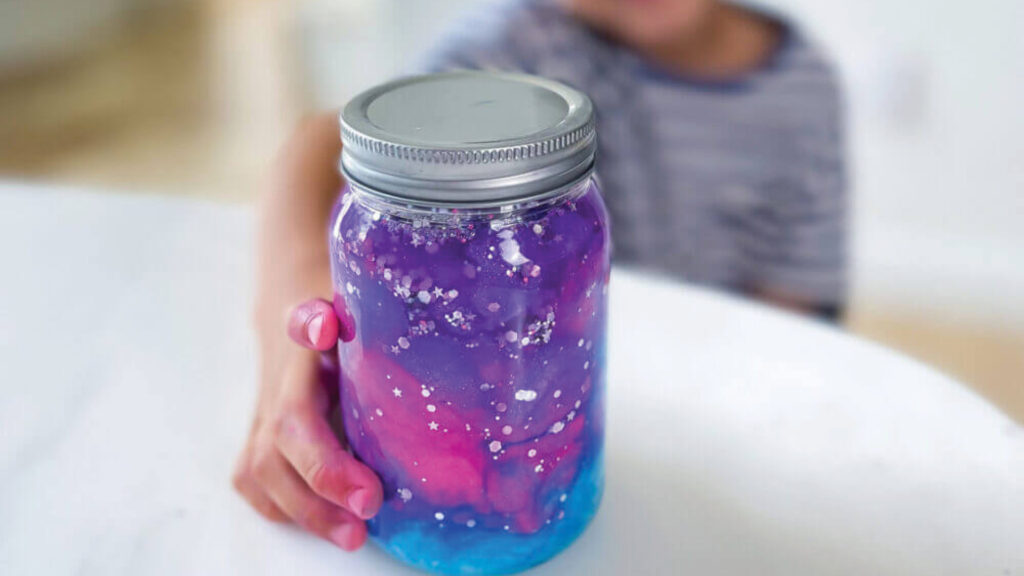 A child holds a jar filled with colorful, glittery layers resembling a galaxy.