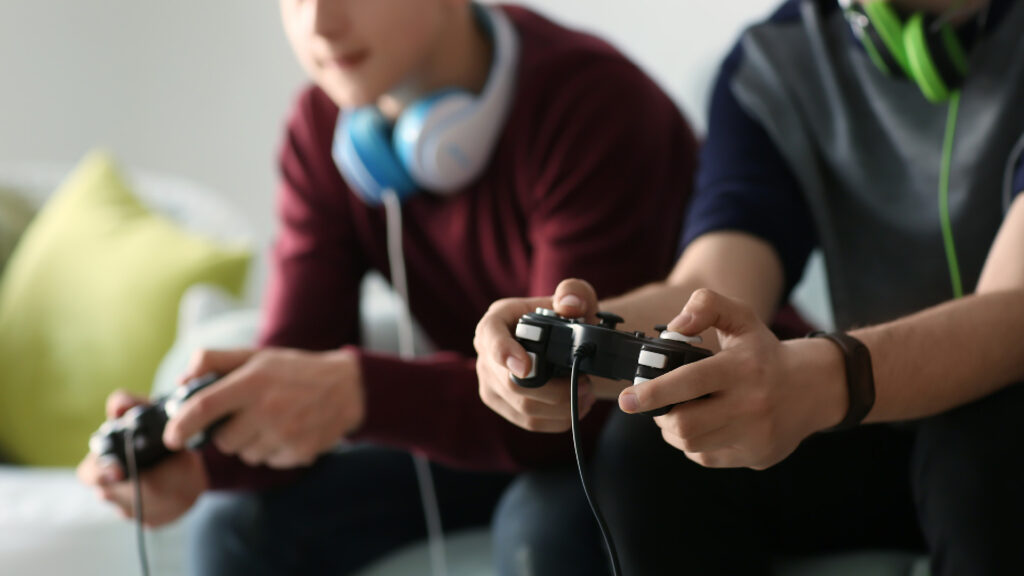 Two people sit closely, focused on playing video games with controllers, while wearing headphones around their necks.
