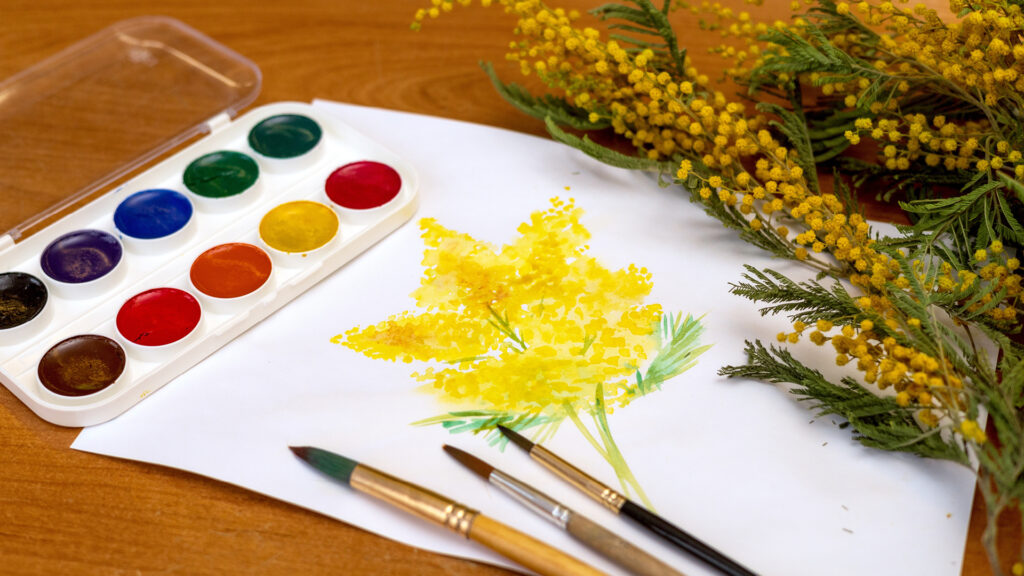 A watercolor painting of yellow flowers on paper, with paints, brushes, and real yellow flowers beside it.