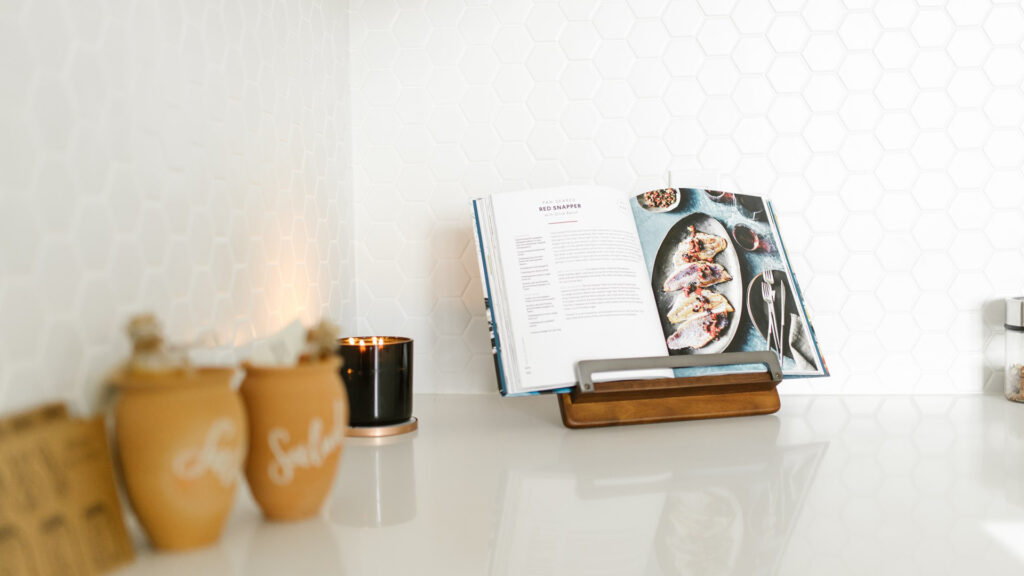 Open cookbook on a stand in a modern kitchen with a honeycomb tile backsplash and various kitchen items nearby.