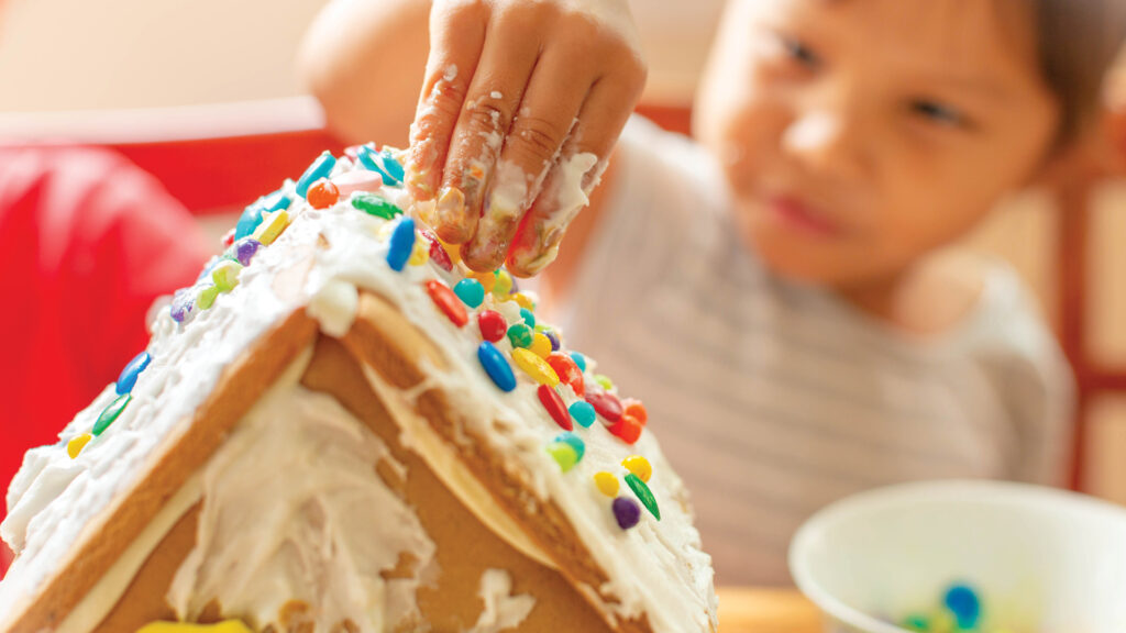Child decorating a gingerbread house with colorful candies and white icing, focusing on their hand.