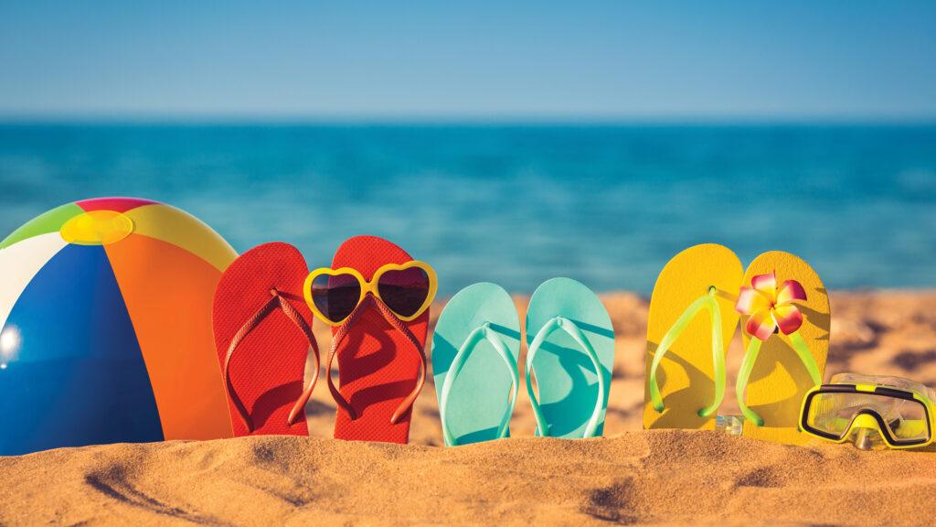 Three pairs of flip-flops, a beach ball, sunglasses, and a snorkel on sand by the ocean.