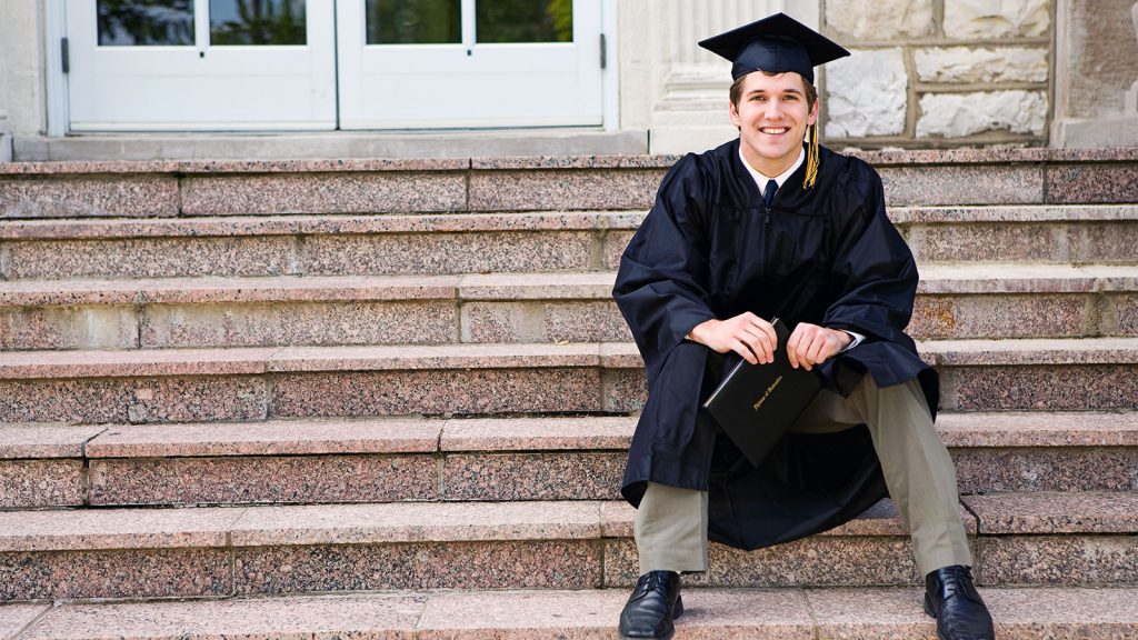 A graduate in cap and gown sits on stone steps holding a diploma, smiling.