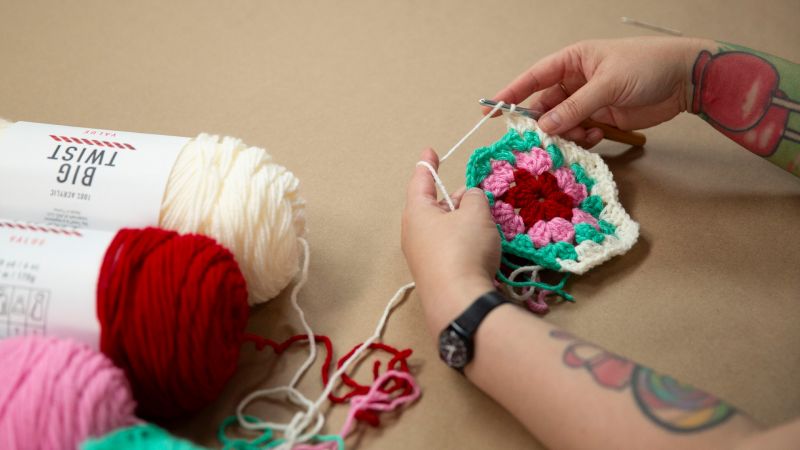 A person with colorful tattoos crochets a multicolored square next to balls of yarn on a beige surface.
