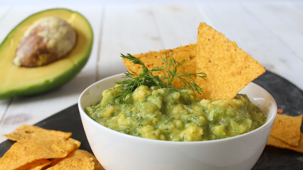 A bowl of guacamole with tortilla chips, garnished with herbs, and a halved avocado in the background.