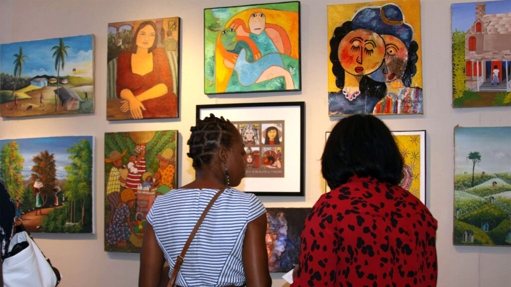 Two people viewing colorful paintings in an art gallery. Several vibrant, diverse artworks are displayed on the wall.