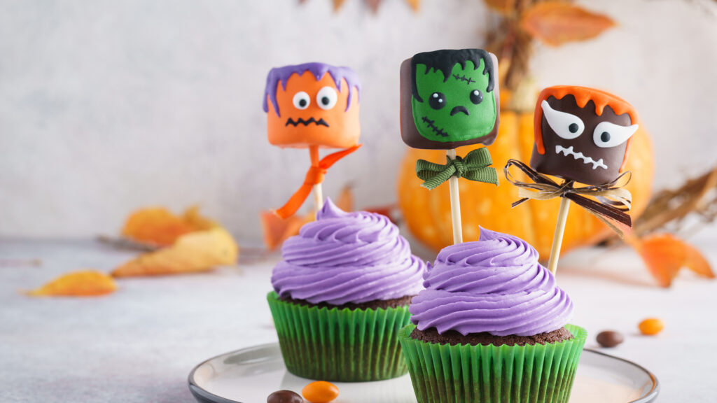 Purple frosted cupcakes with Halloween monster marshmallow toppers, pumpkins, and autumn leaves in background.