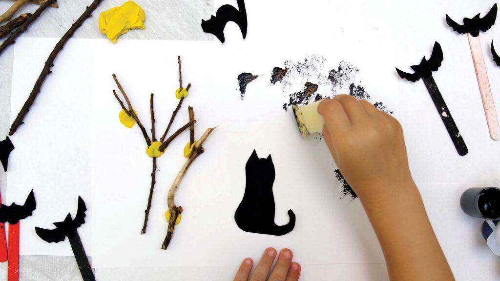 A child makes Halloween art with sticks, yellow clay, and black cat and bat cutouts on white paper.