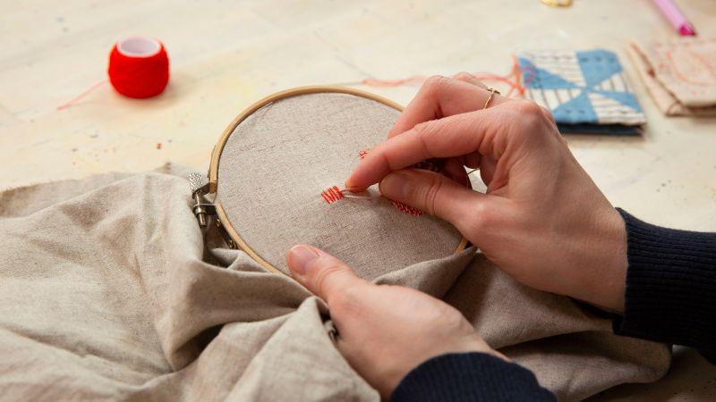 A person stitching red thread onto fabric held in an embroidery hoop, with sewing materials nearby.