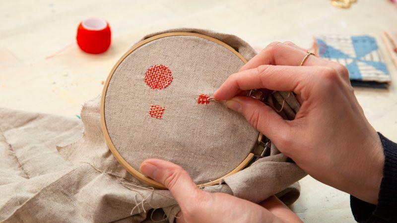 Hands embroidering red patterns on beige fabric in a wooden hoop, with thread and needle visible.