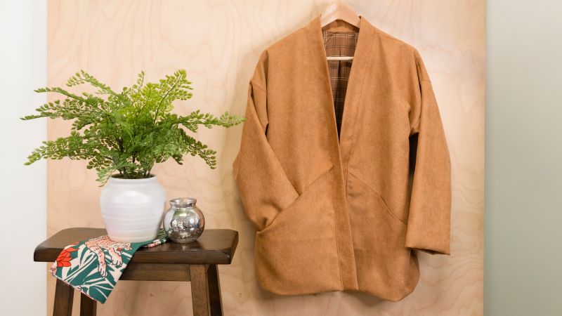 A brown cardigan on a hanger next to a potted plant and a small silver jar on a wooden stool.