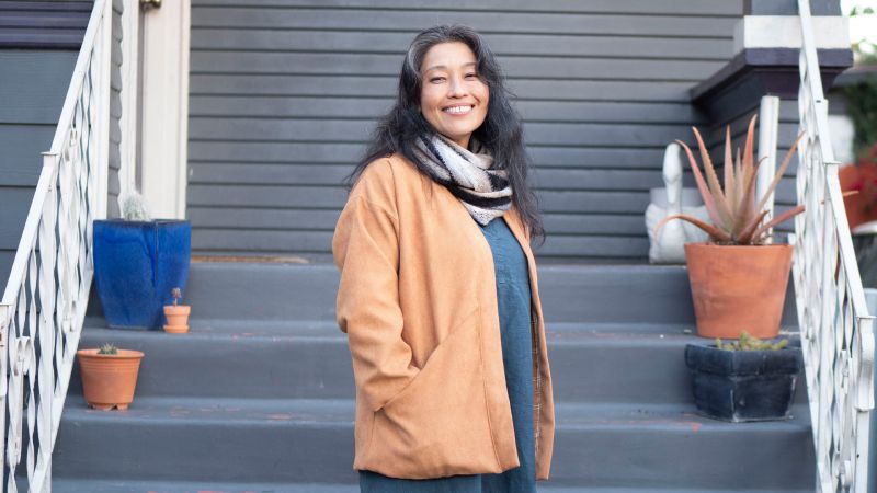 A smiling person stands on steps wearing a brown coat and scarf, with potted plants on both sides of the steps.