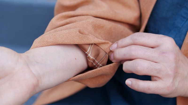 Person rolling up the sleeve of an orange shirt, revealing delicate bracelets on their wrist.