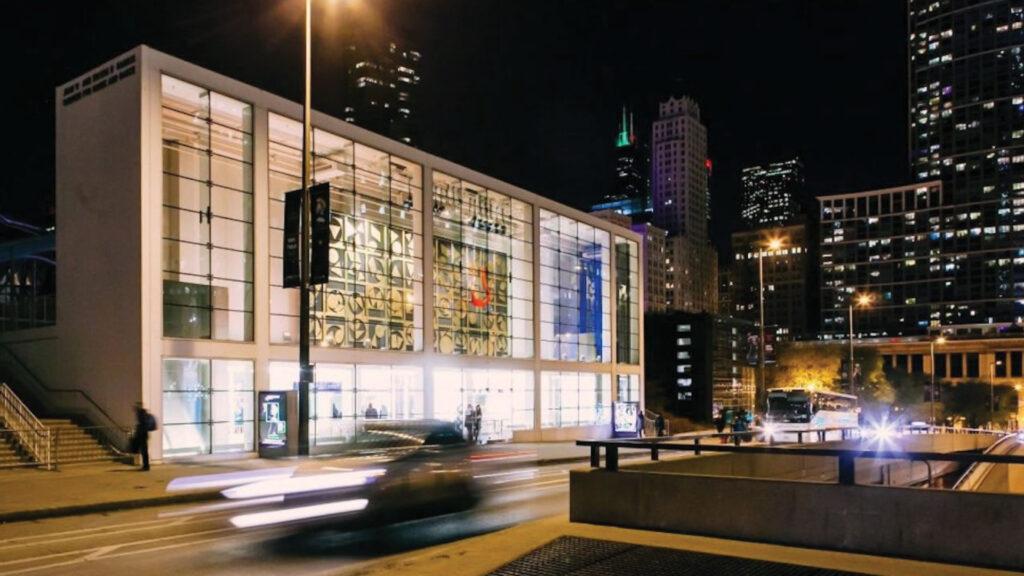 Modern glass building lit at night, with cars driving past in the foreground and city skyline in the background.