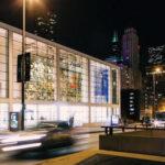 Modern glass building lit at night, with cars driving past in the foreground and city skyline in the background.