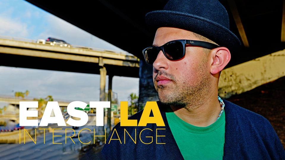 A man in a hat and sunglasses stands under a freeway overpass with "East LA Interchange" text beside him.