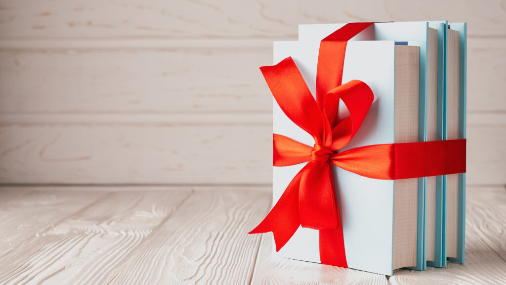 Three books wrapped together with a red ribbon bow, placed on a light wooden surface.