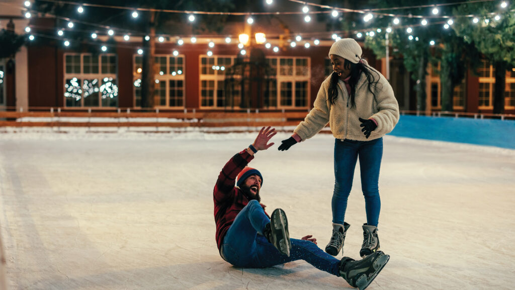Two people ice skating outdoors at night; one has fallen and the other is helping them up, both are smiling.