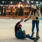 Two people ice skating outdoors at night; one has fallen and the other is helping them up, both are smiling.