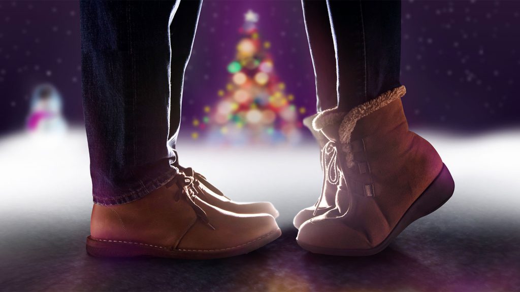 Two people wearing cozy boots stand on tiptoes to kiss under snowy night with a blurred Christmas tree in the background.