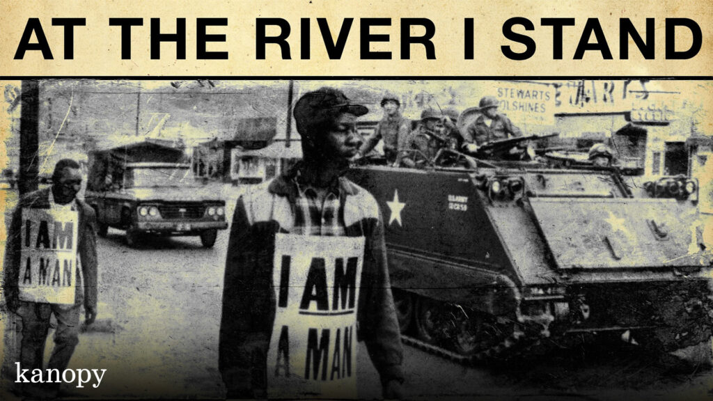 Black-and-white photo of a civil rights protester wearing an "I AM A MAN" sign with military vehicles behind.