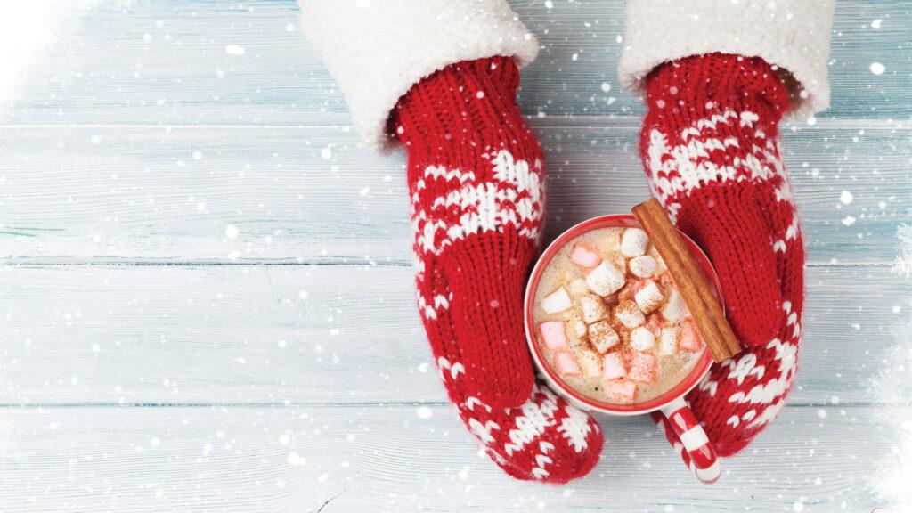 Hands in red mittens holding a cup of hot chocolate with marshmallows and a cinnamon stick, in a snowy setting.