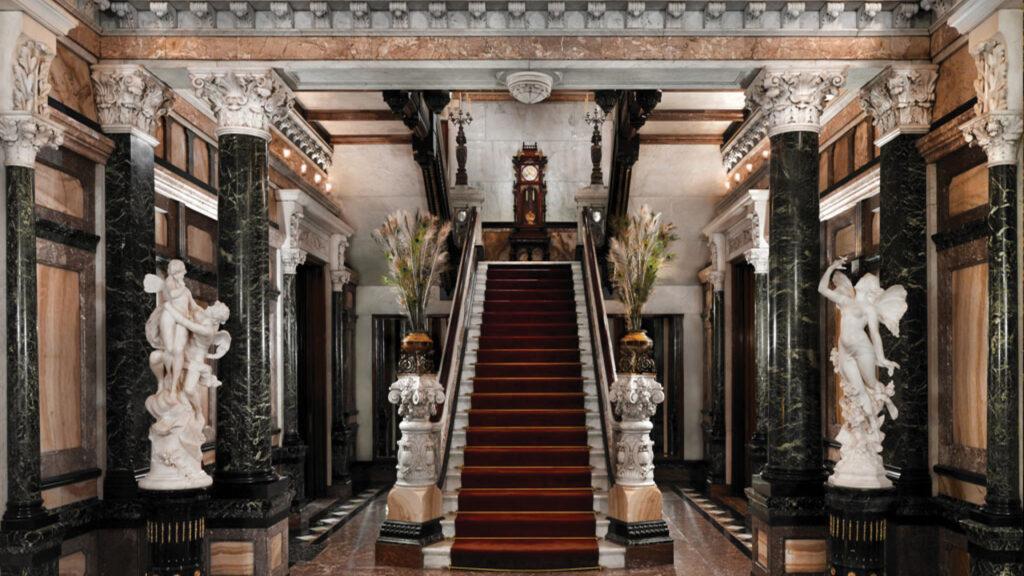 Opulent marble staircase with red carpet, ornate pillars, statues, and floral arrangements in a grand foyer.