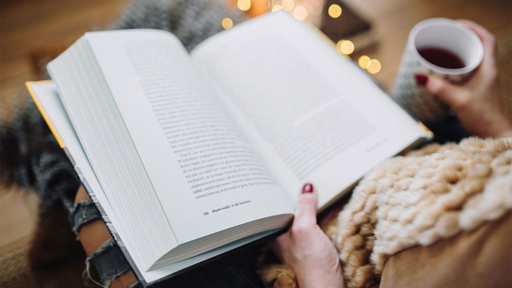 Person reading a book and holding a cup of tea, with cozy, warm lighting in the background.