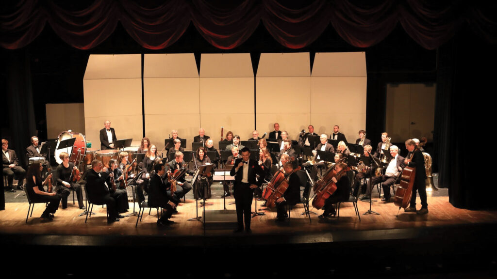 An orchestra performing on stage with musicians playing various instruments, including strings, brass, and woodwinds.