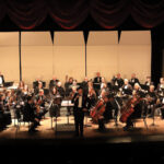 An orchestra performing on stage with musicians playing various instruments, including strings, brass, and woodwinds.