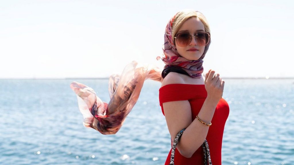 A woman in a red dress with a headscarf and sunglasses stands in front of a body of water on a sunny day.