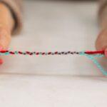Hands with pink nail polish hold a twisted strand of red and blue yarn on a light surface.