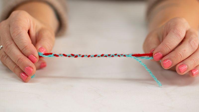 Hands with pink nail polish hold a twisted strand of red and blue yarn on a light surface.