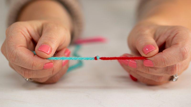 Close-up of hands holding two pieces of yarn, one blue and one red, joined together with a knot.