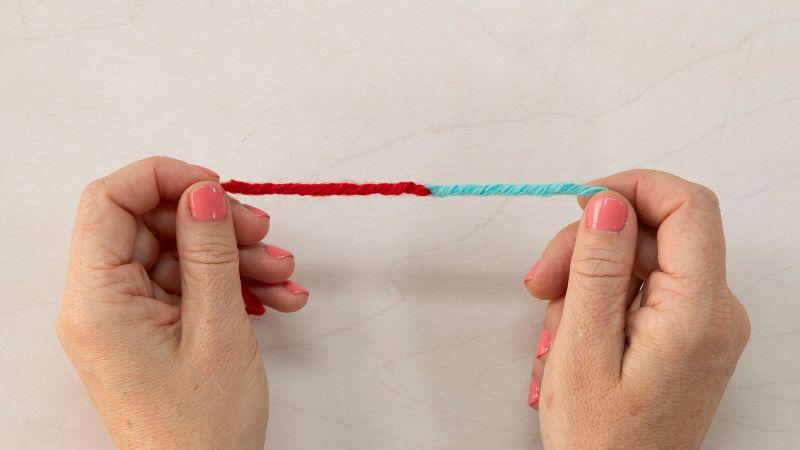 Two hands holding a joined strand of red and blue yarn against a light background.