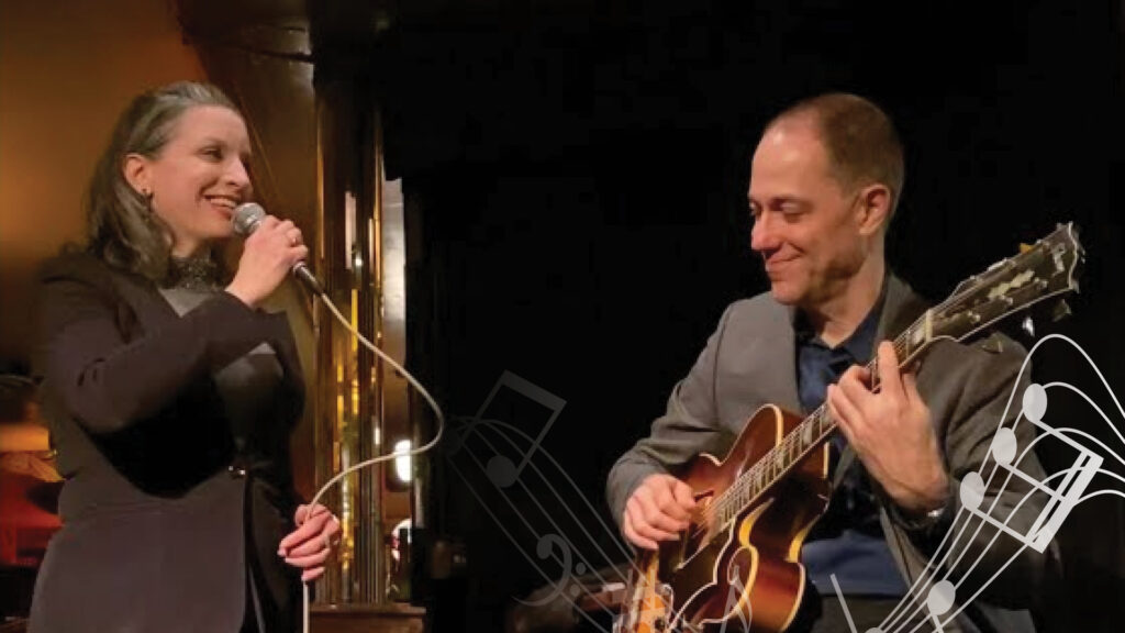 A woman sings into a microphone while a man plays guitar beside her on stage, both smiling.