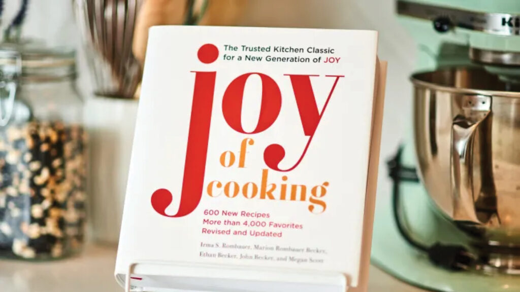 Joy of Cooking" cookbook displayed on a stand in a kitchen next to a mixer and jars.