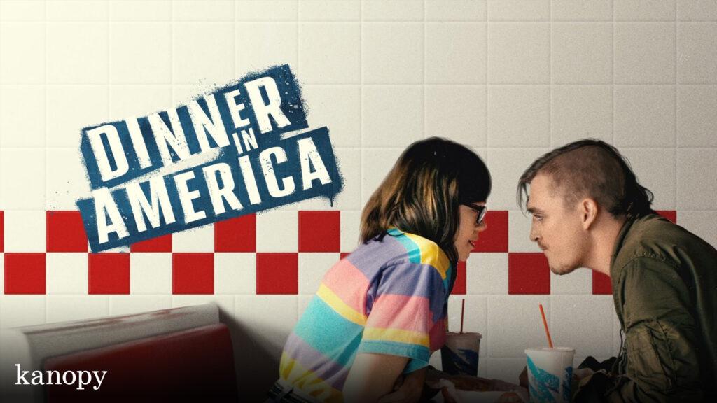 A couple sits close together in a diner booth with "Dinner in America" text on a tiled wall behind them.