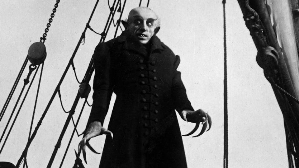 A pale figure with long claws and sharp teeth stands menacingly on a ship's deck, dressed in dark, vintage clothing.