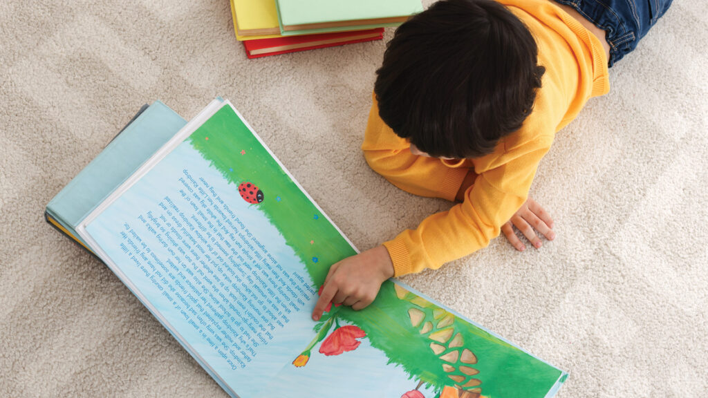 Child in a yellow sweater lies on carpet, reading and pointing at a colorful picture book with flowers and a ladybug.