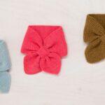 Three knitted bow-tie scarves in light blue, pink, and brown on a light wooden surface with knitting needles.