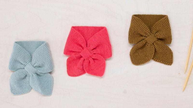 Three knitted bow-tie scarves in light blue, pink, and brown on a light wooden surface with knitting needles.